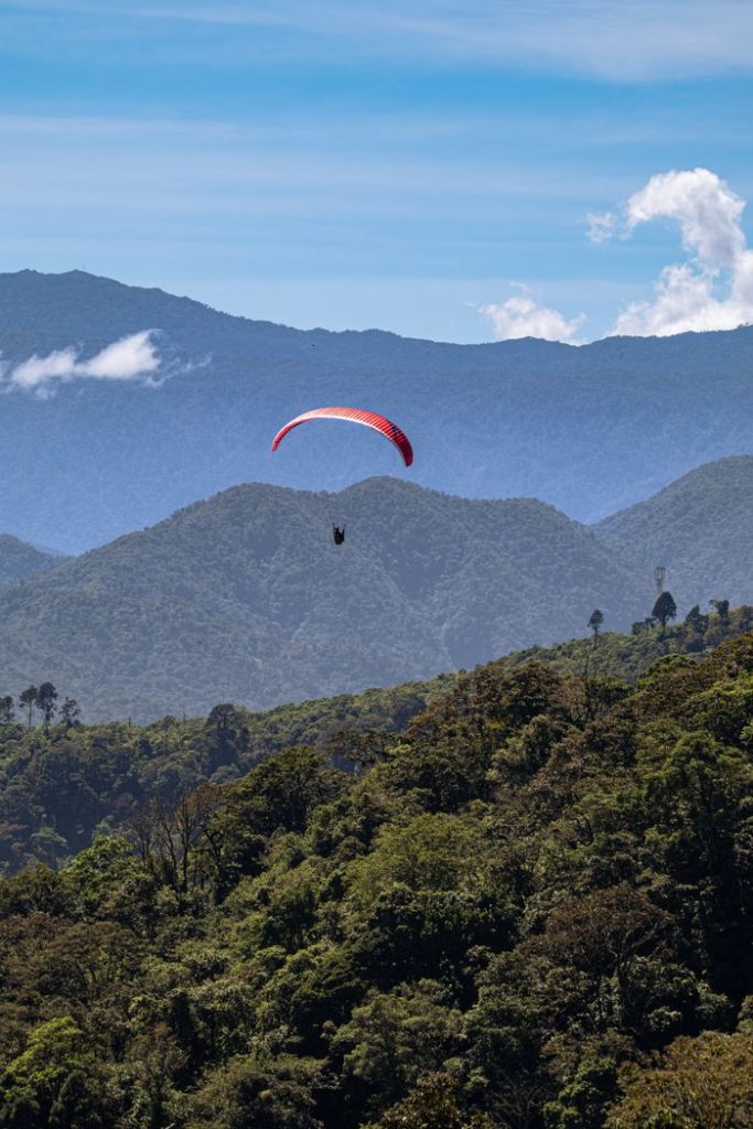 A paraglider soars above vibrant green landscapes and distant mountain ranges under a clear blue sky.